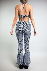 A woman wearing a black and white geometric printed jumpsuit with bell bottoms. The jumpsuit is a halter neck with cutouts on either side and a tie back. The woman is facing away from the camera.