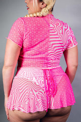 A woman wearing a mesh cropped pink and white t shirt and matching skirt. She is facing away from the camera.