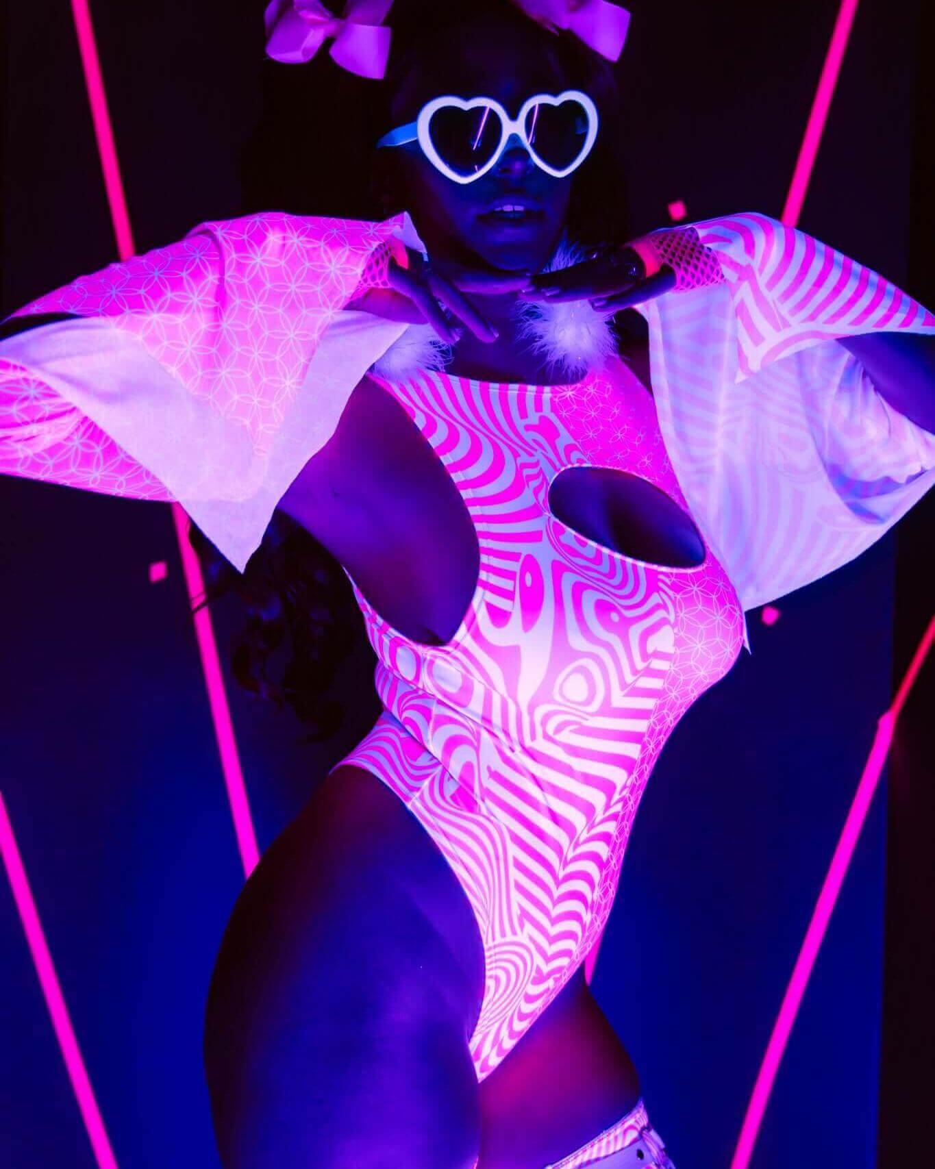 Model wearing neon rave outfit with heart-shaped glasses, glowing under UV light, showcasing vibrant colors and patterns.