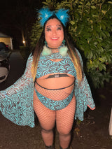 raver wears blue and black festival outfit with buckle details and fishnets in front of green trees