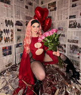 Woman in a red cut-out bodysuit with pink hair and heart balloons poses amidst newspaper-clad walls and shiny foil.