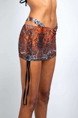 Profile view of a model wearing a brown sheer mini skirt with side straps from Freedom Rave Wear, highlighting the detailed pattern.