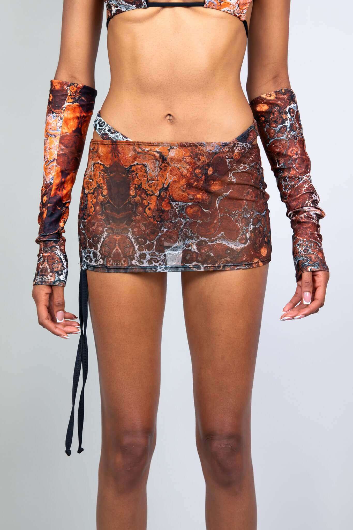 Front view of a model in a brown sheer mini skirt with matching arm sleeves from Freedom Rave Wear, highlighting the detailed pattern.