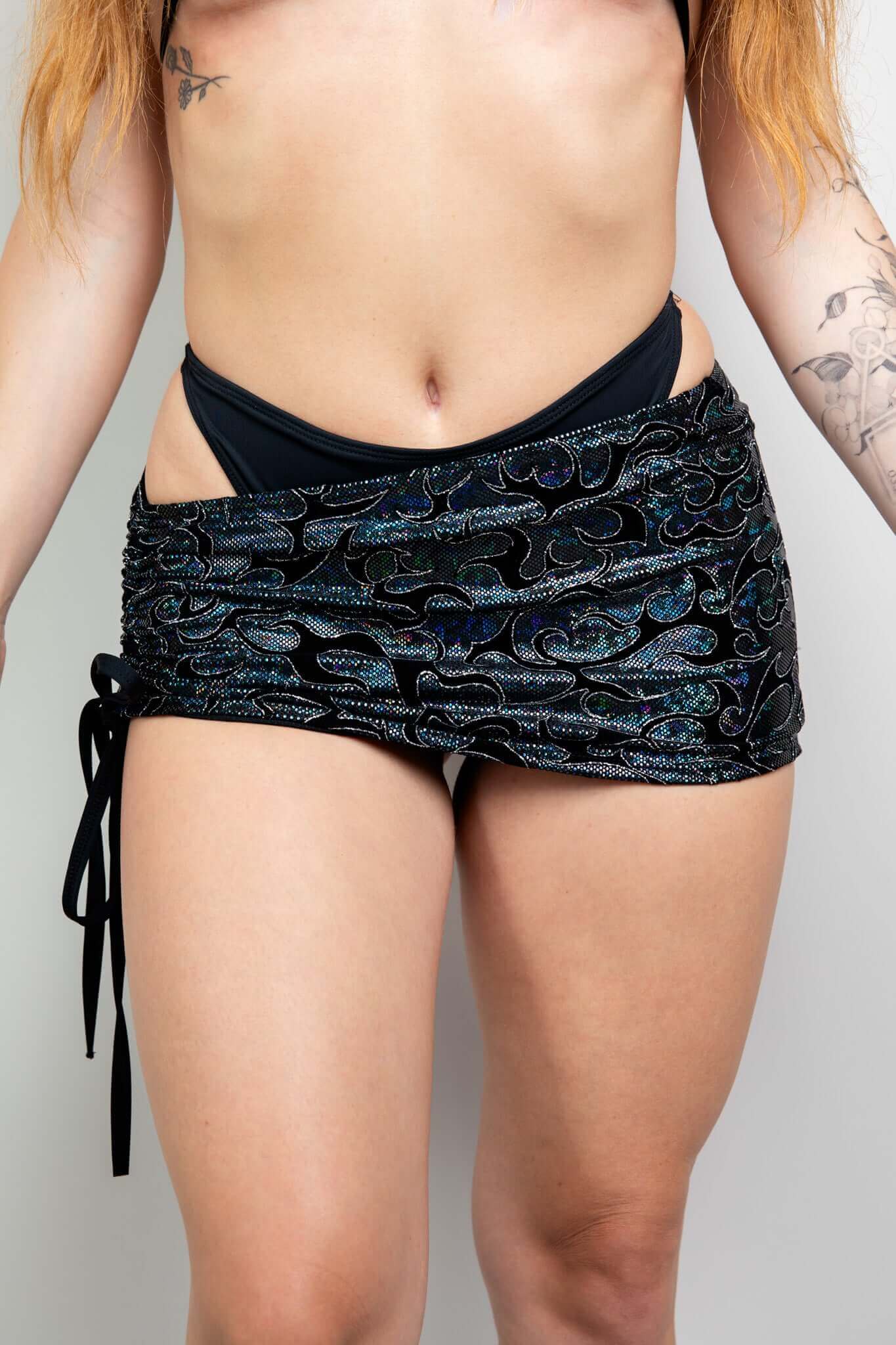 Model wearing a black holographic mini skirt with side tie detail by Freedom Rave Wear, showcasing intricate patterns and body tattoos.