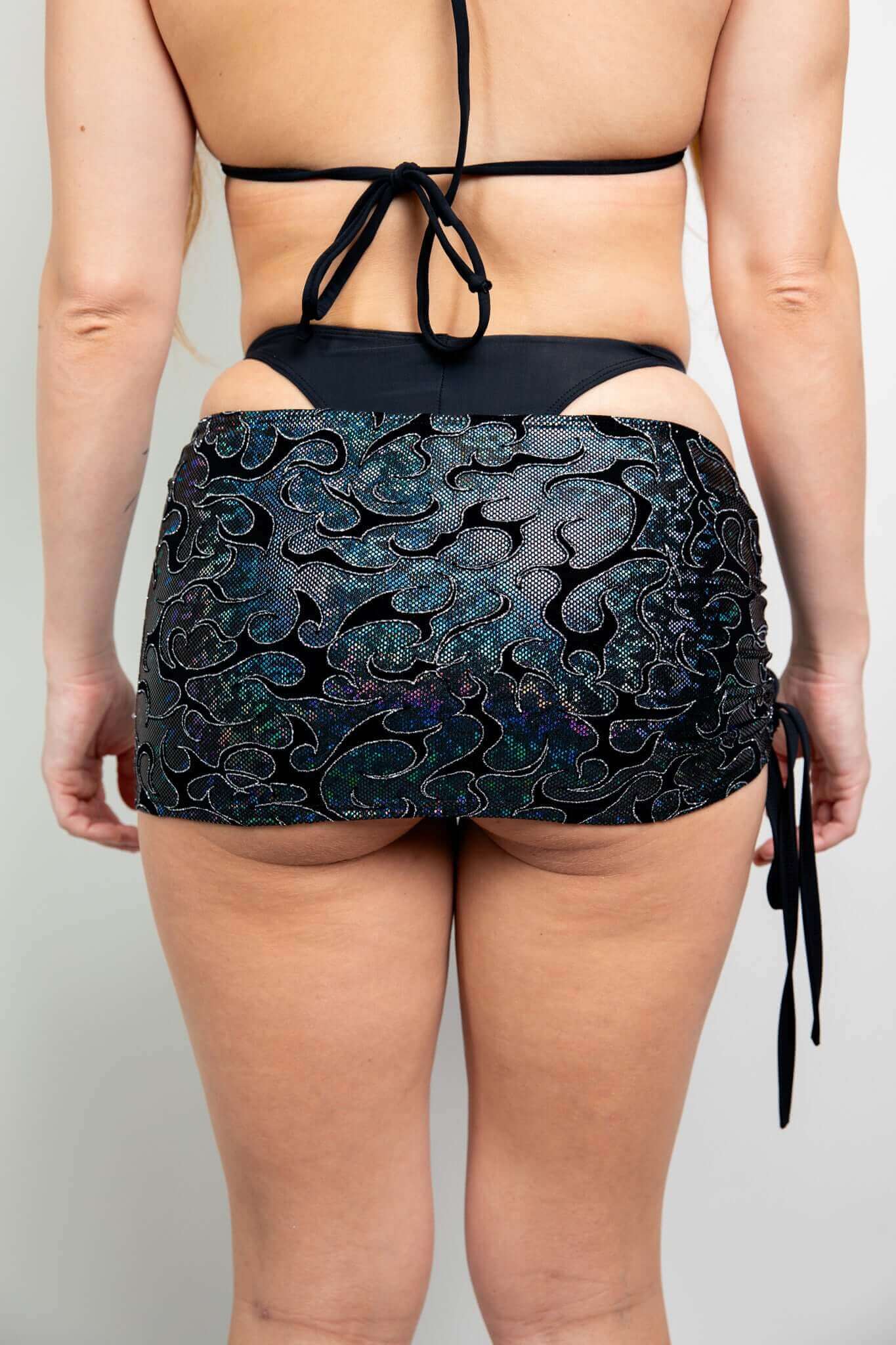 Rear view of black holographic mini skirt by Freedom Rave Wear, featuring side tie and swirling patterns, perfect for rave wear.