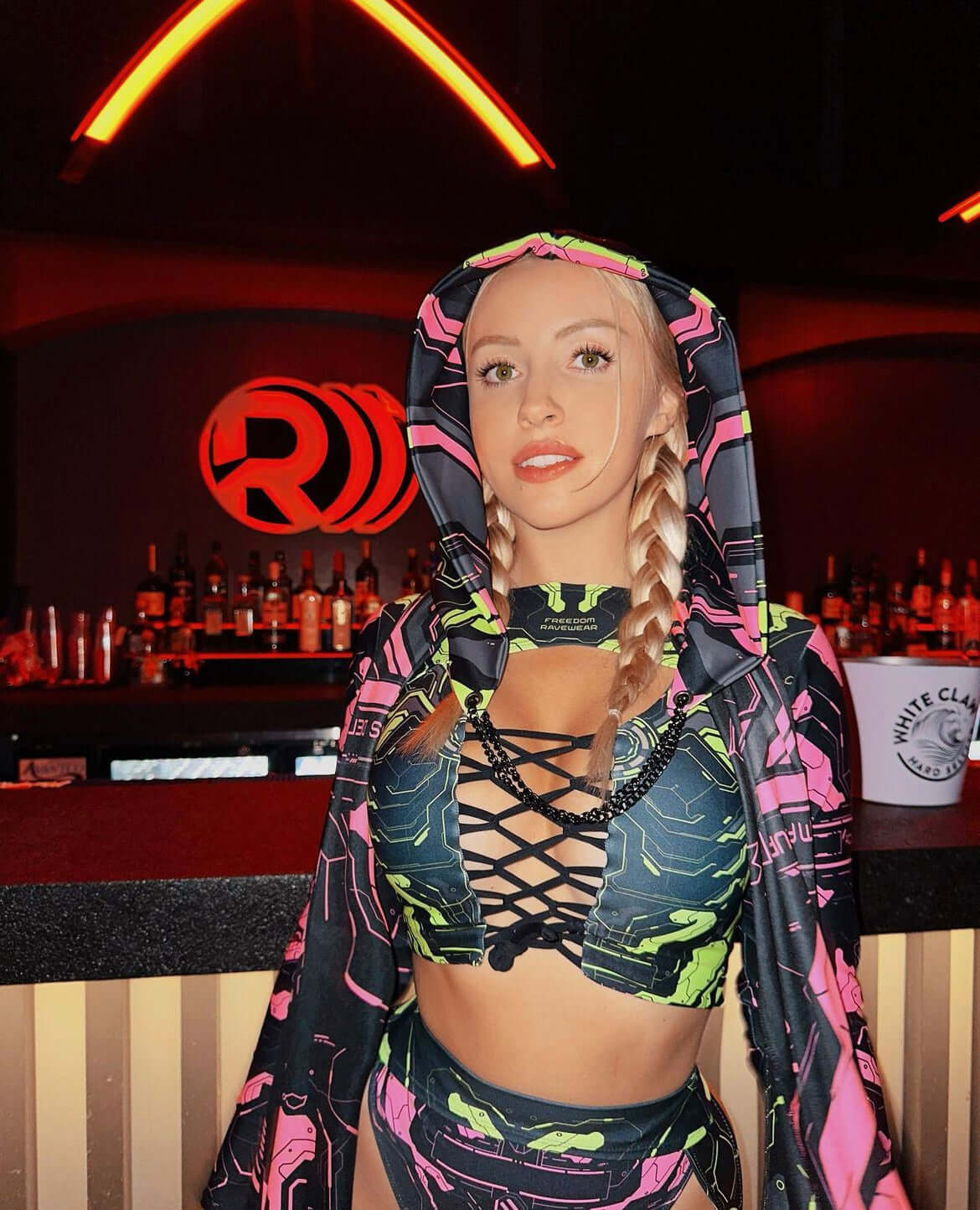 Model in a neon cyber rave outfit poses playfully at a bar with striking red neon lights in the background.