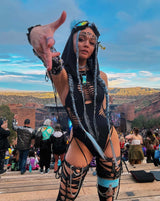 Confident festival enthusiast in a dynamic rave outfit, extending a hand towards the camera with a scenic concert backdrop, adorned in a black braided bodysuit and blue hair accents