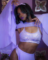 Poc rave girl poses with a mystical aura in a sparkling lilac rave set, complemented by butterfly artwork backdrop