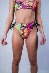 Model wearing Intensity Lace Up Brazilian Bottoms in vibrant patterns, ideal for rave outfits and stylish summer swimwear.