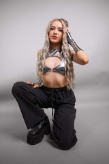 A girl kneeling in front of a blank background. She is wearing a silver crop top and black, loose fitting pants.