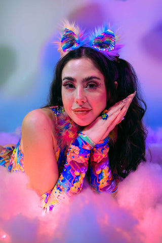 A woman laying on a cloud wearing a rainbow outfit and rainbow sequin ears.