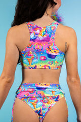 An up close photo of a woman wearing a rainbow crop top with whimsical characters on it and matching bikini bottoms. She is facing away from the camera.