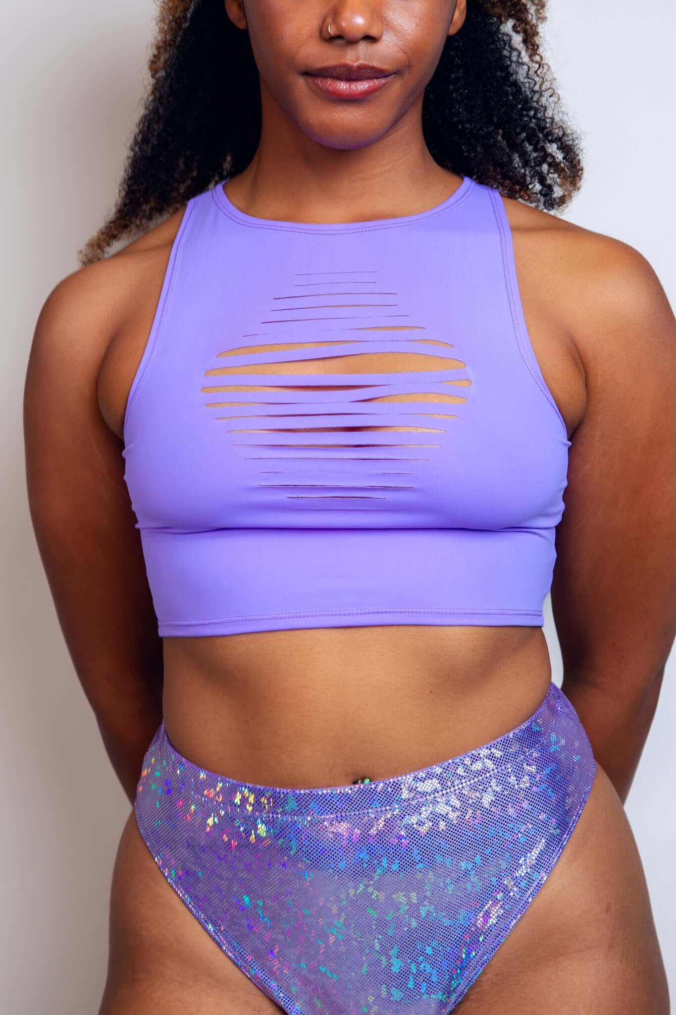 Confident woman in a lavender Freedom Rave Wear crop top with a distinctive front slit design and gold details, paired with a shimmering purple high-waist rave bottom