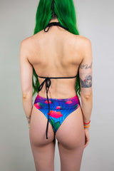 Rear view of a model in Freedom Rave Wear, featuring a multicolored high-waisted thong and black tie-up halter top.