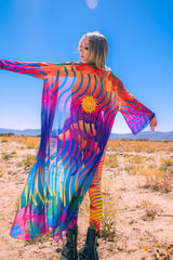 Freedom Rave Wear flowing robe in vibrant desert scene, featuring sun and wave motifs, modeled elegantly.