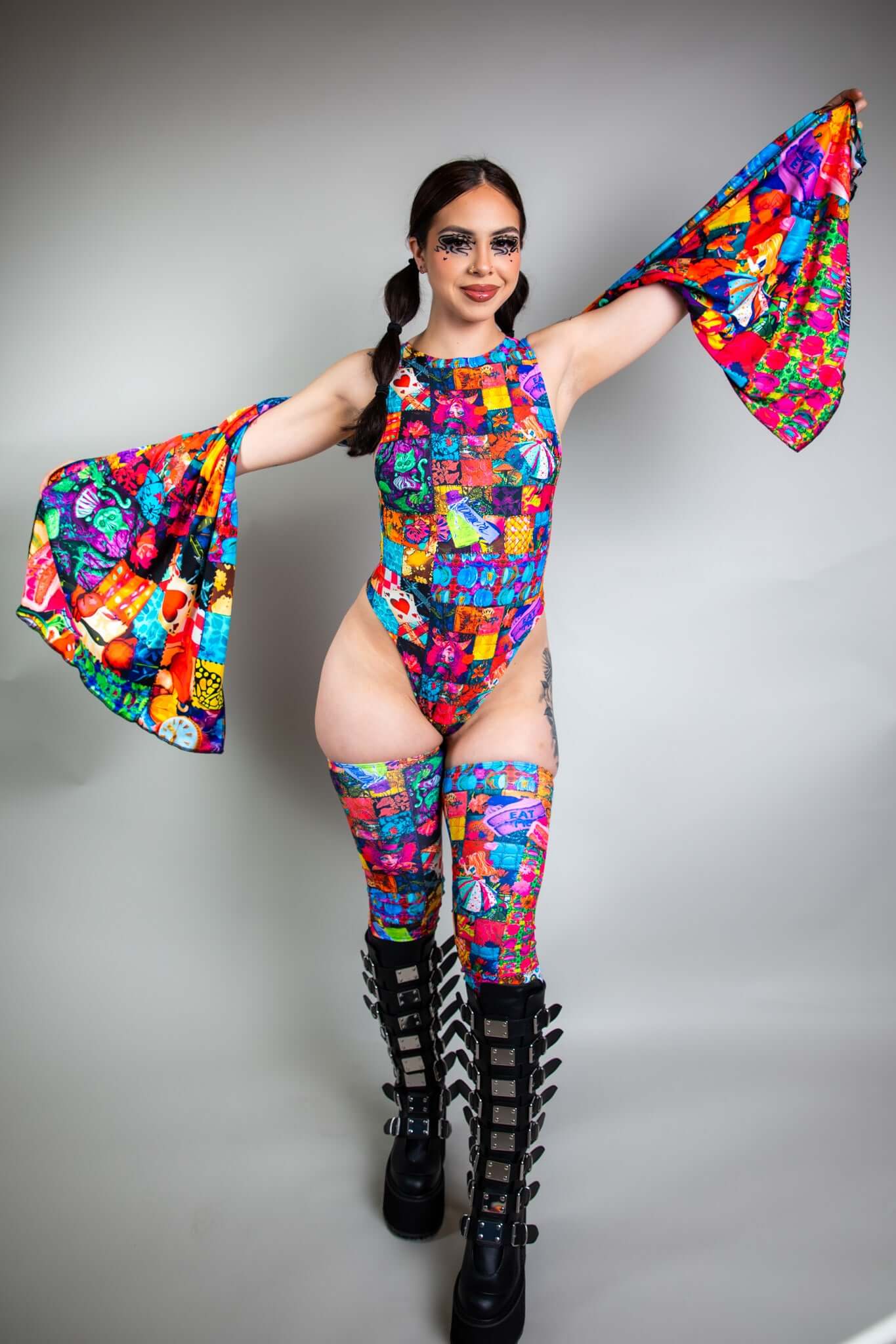 A girl with swirly black eye makeup wearing a rainbow patchwork bodysuit with a matching pashmina draped over her arms.