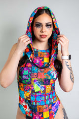 A girl with black swirly eye makeup wearing a rainbow patchwork hood and matching bodysuit.