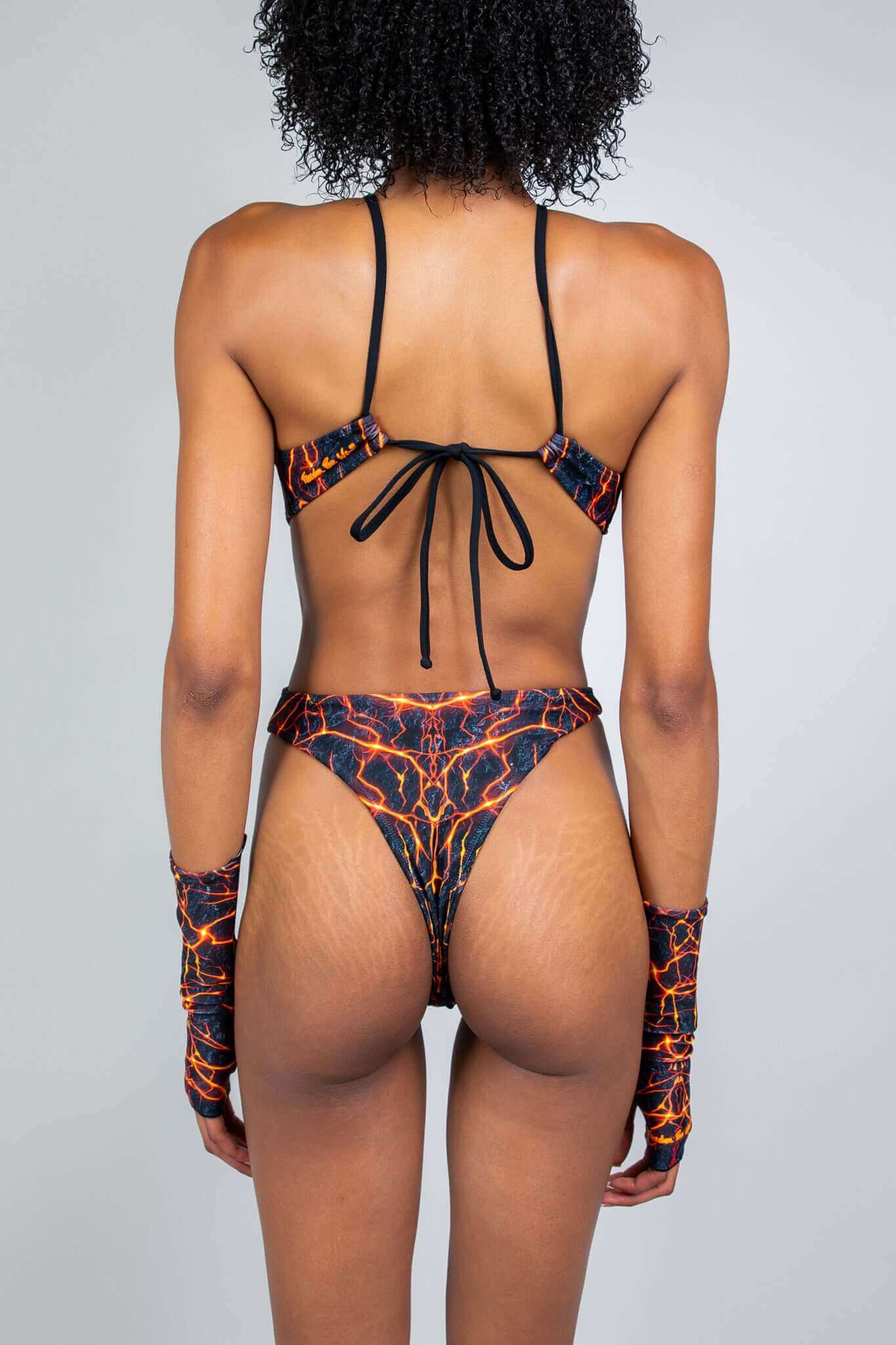 Model wearing a Magma Halo Bodysuit with matching gloves, back view featuring a strappy design, red and black magma print, and thong-cut bottom.