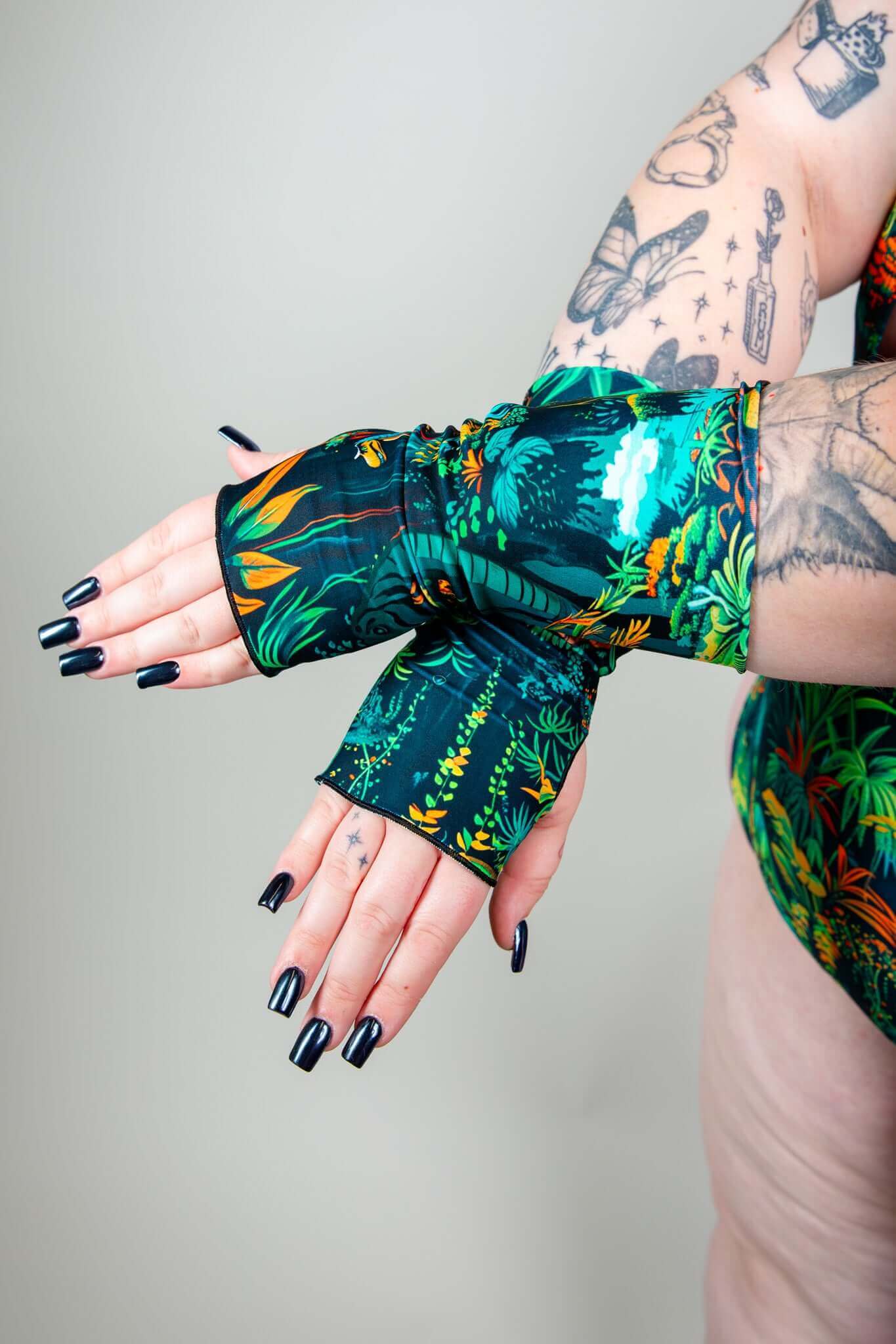 Close-up of hands wearing Freedom Rave Wear's tropical dinosaur print rave gloves, featuring vibrant colors and paired with black nail polish and tattoos.