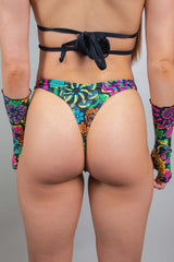 Rear view of a model in Freedom Rave Wear high-waisted rave bottoms, featuring a rich psychedelic pattern. The design highlights intricate floral motifs in a spectrum of vibrant colors, perfectly paired with a black crisscross top and arm accessories for a festival-ready look.