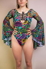 Front view of a confident model in a Freedom Rave Wear Goddess bodysuit featuring an intricate lace-up detail, psychedelic floral print, and dramatic bell sleeves.