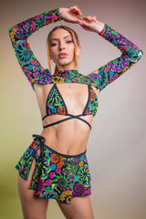 Model striking a pose in a Freedom Rave Wear bikini top with spectra sleeves and skirt, highlighted by a bold, colorful neon floral print against a warm backdrop.