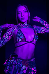 Model in a Freedom Rave Wear floral bikini top and skirt set with spectra sleeves, bathed in purple light that casts a glow on the unique rave attire.