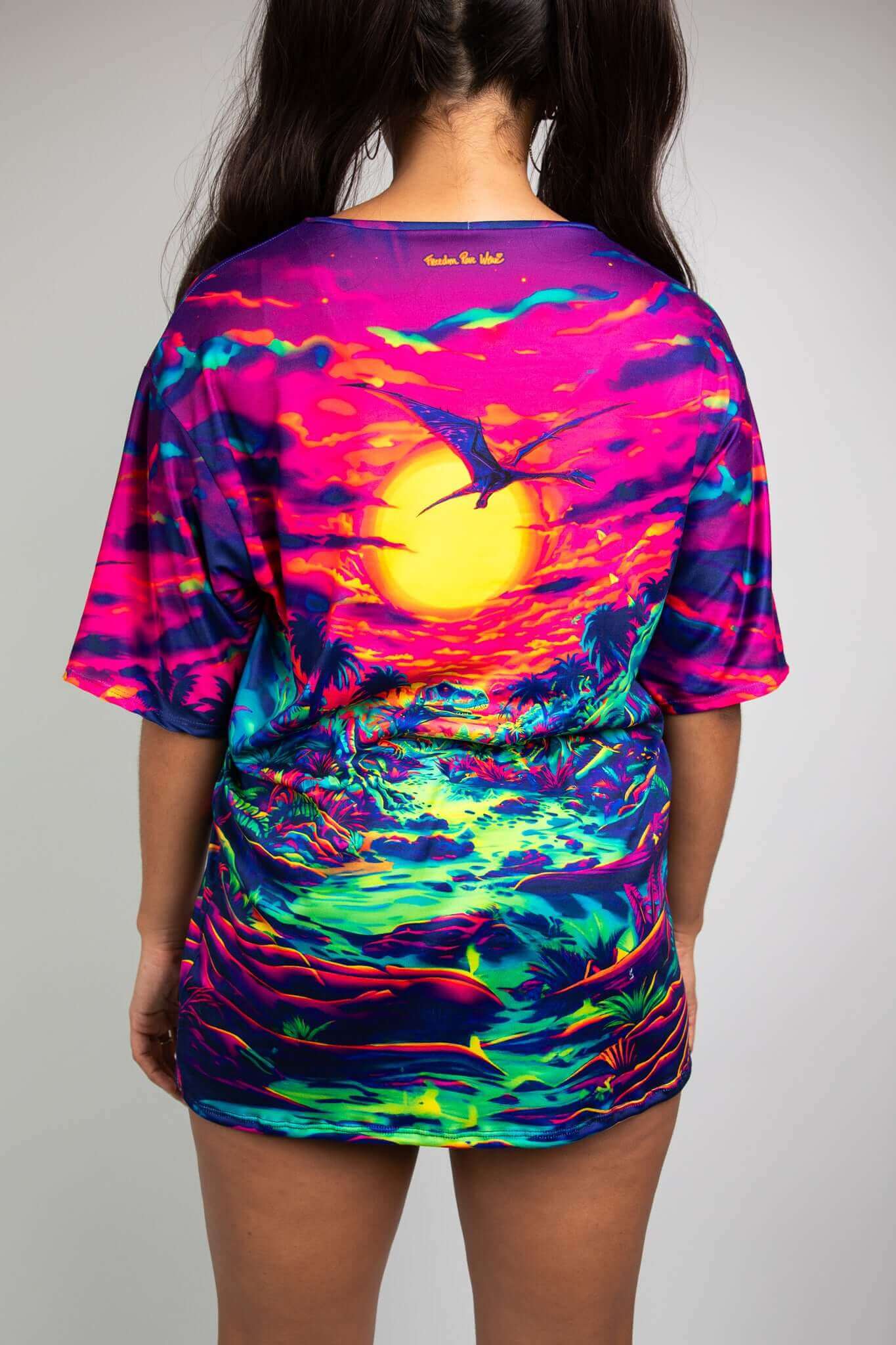 Model wearing Freedom Rave Wear t-shirt from the back, showcasing a vivid dinosaur scene under a neon sunset for bold rave style.