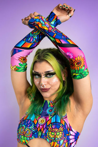 A photo of a girl with green hair holding her arms in an x above her head. She is wearing stained glass printed arm sleeves and a matching top.