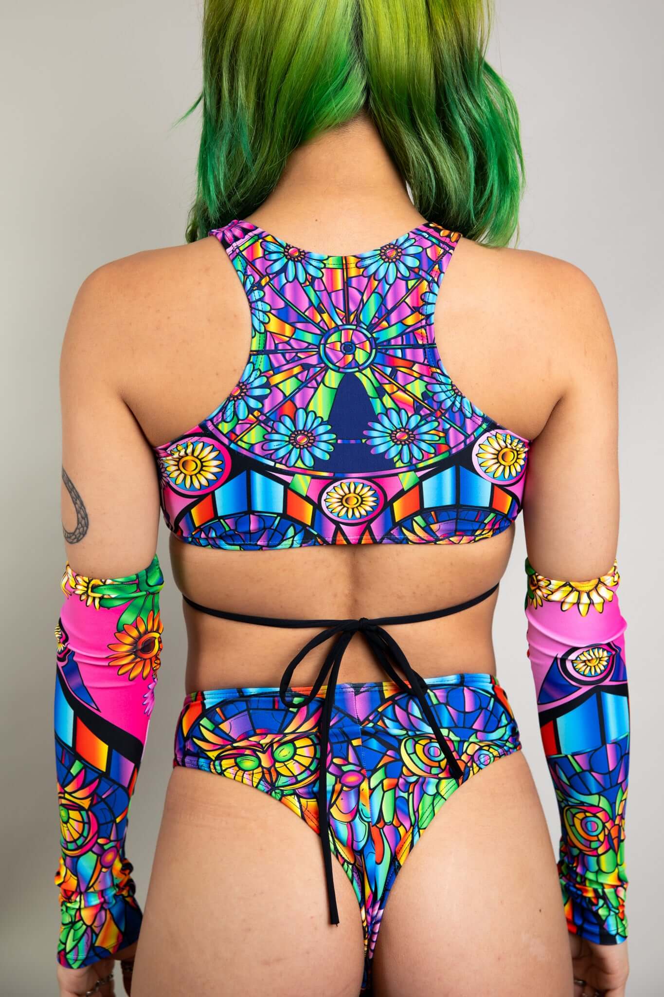 An up close photo of a girl wearing a rainbow stained glass printed crop top with black ties. She is facing away from the camera.