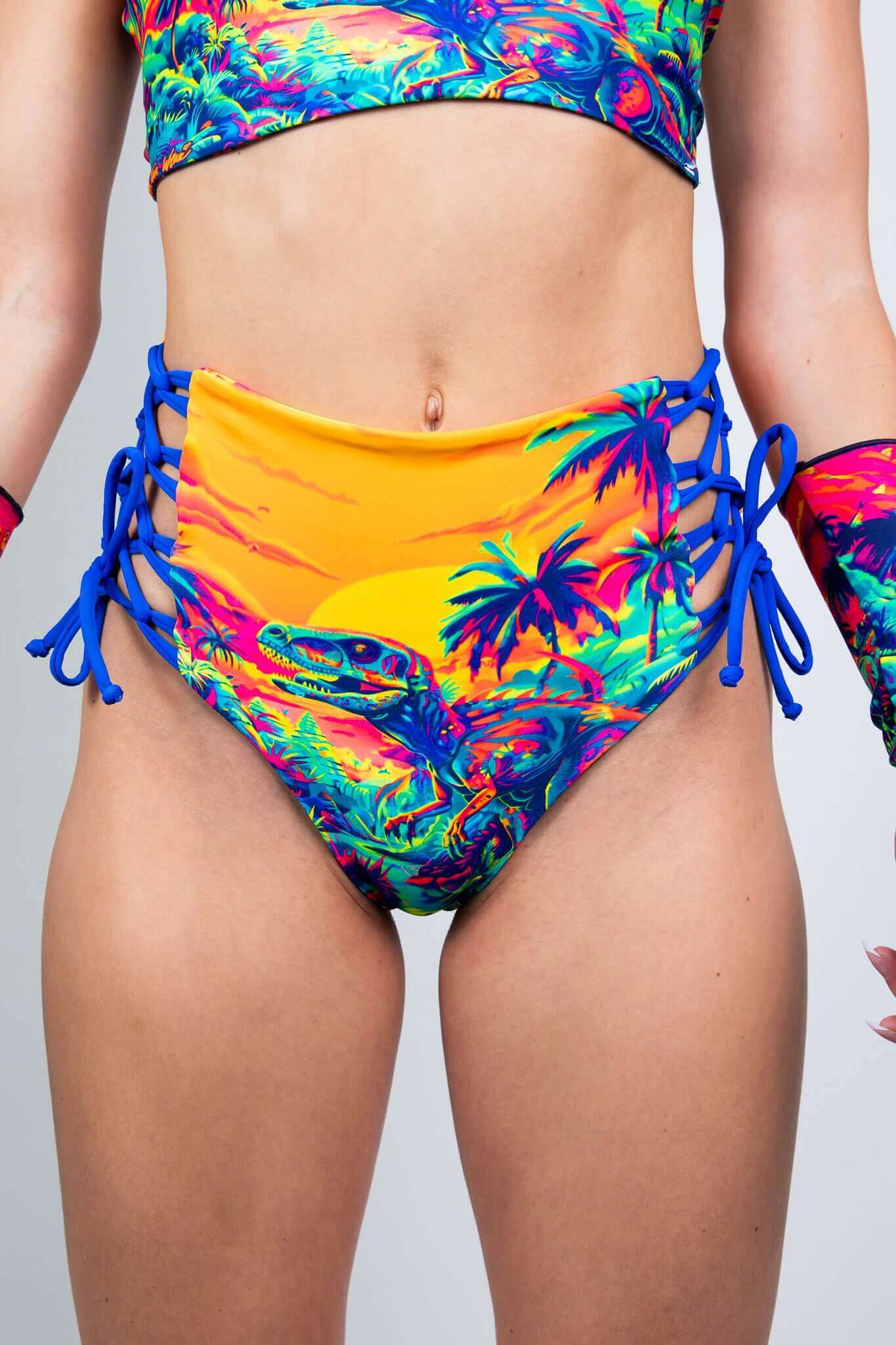 Model showcasing Freedom Rave Wear's high-waisted rave shorts with vibrant dinosaur sunset print and blue lace-up sides, paired with matching gloves.