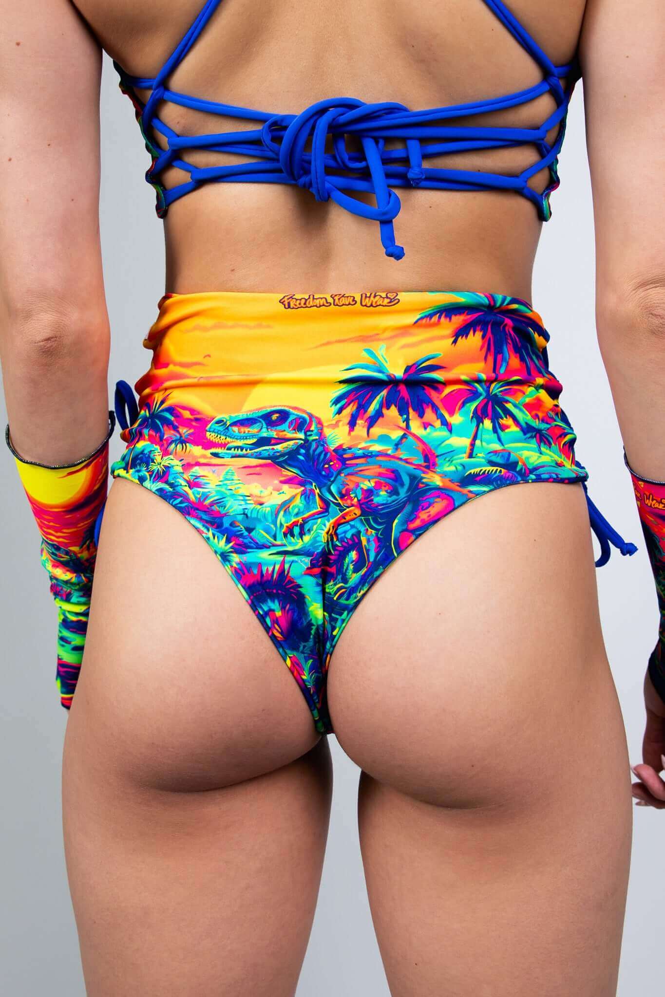 Back view of a model in Freedom Rave Wear's high-waisted rave shorts featuring a vibrant dinosaur sunset print, blue lace-up back detail, and matching arm sleeves.