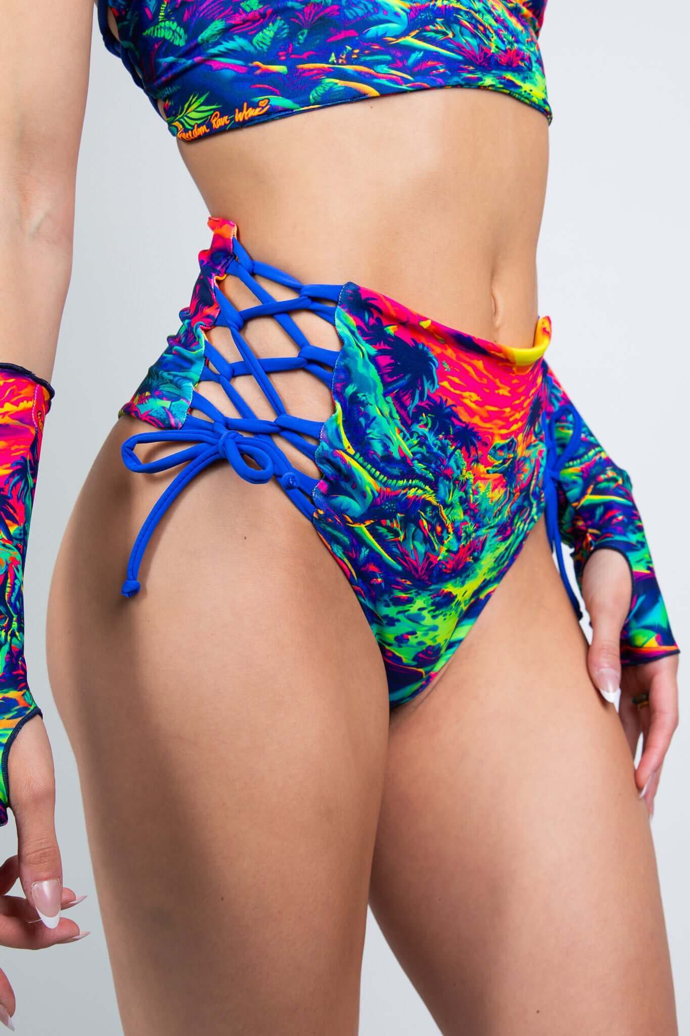 Close-up side view of a model in Freedom Rave Wear's high-waisted rave shorts featuring a vibrant dinosaur sunset print, blue lace-up sides, and matching gloves