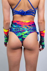 Back view of a model wearing Freedom Rave Wear's high-waisted rave shorts with a vibrant dinosaur sunset print, blue lace-up back detail, and matching arm sleeves.