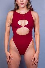 Woman models a scarlet Freedom Rave Wear keyhole rave bodysuit, featuring a bold circular cutout at the chest and a sleek, form-fitting design, set against a soft gray background, enhancing her confident pose.
