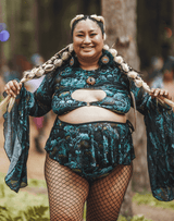 Smiling person wearing a turquoise rave outfit with a cutout top and layered skirt, posing in a forest setting.