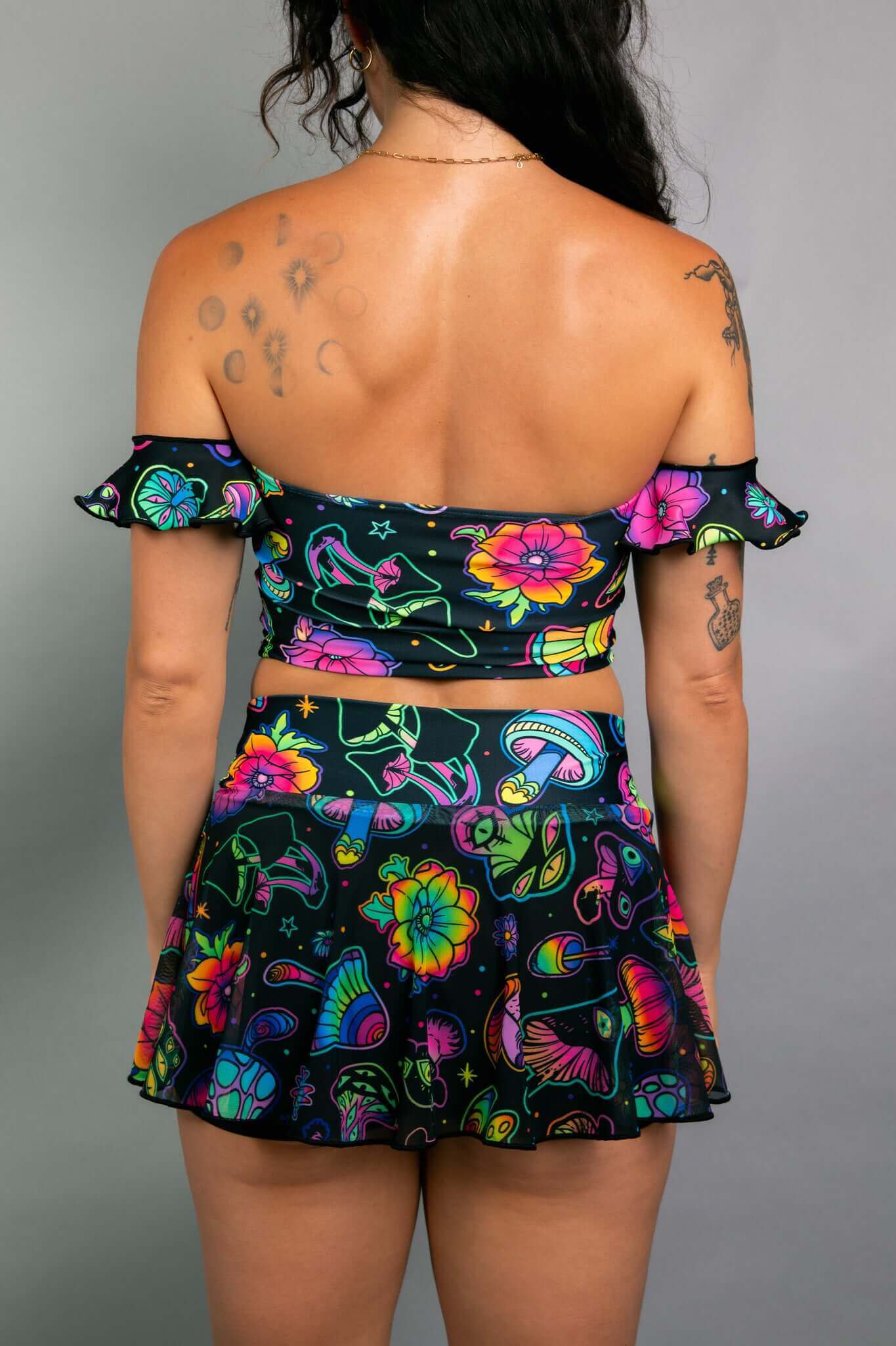 Back view of a model wearing a Freedom Rave Wear crop top and skater skirt set with off-the-shoulder ruffles, featuring a colorful psychedelic mushroom and floral print.