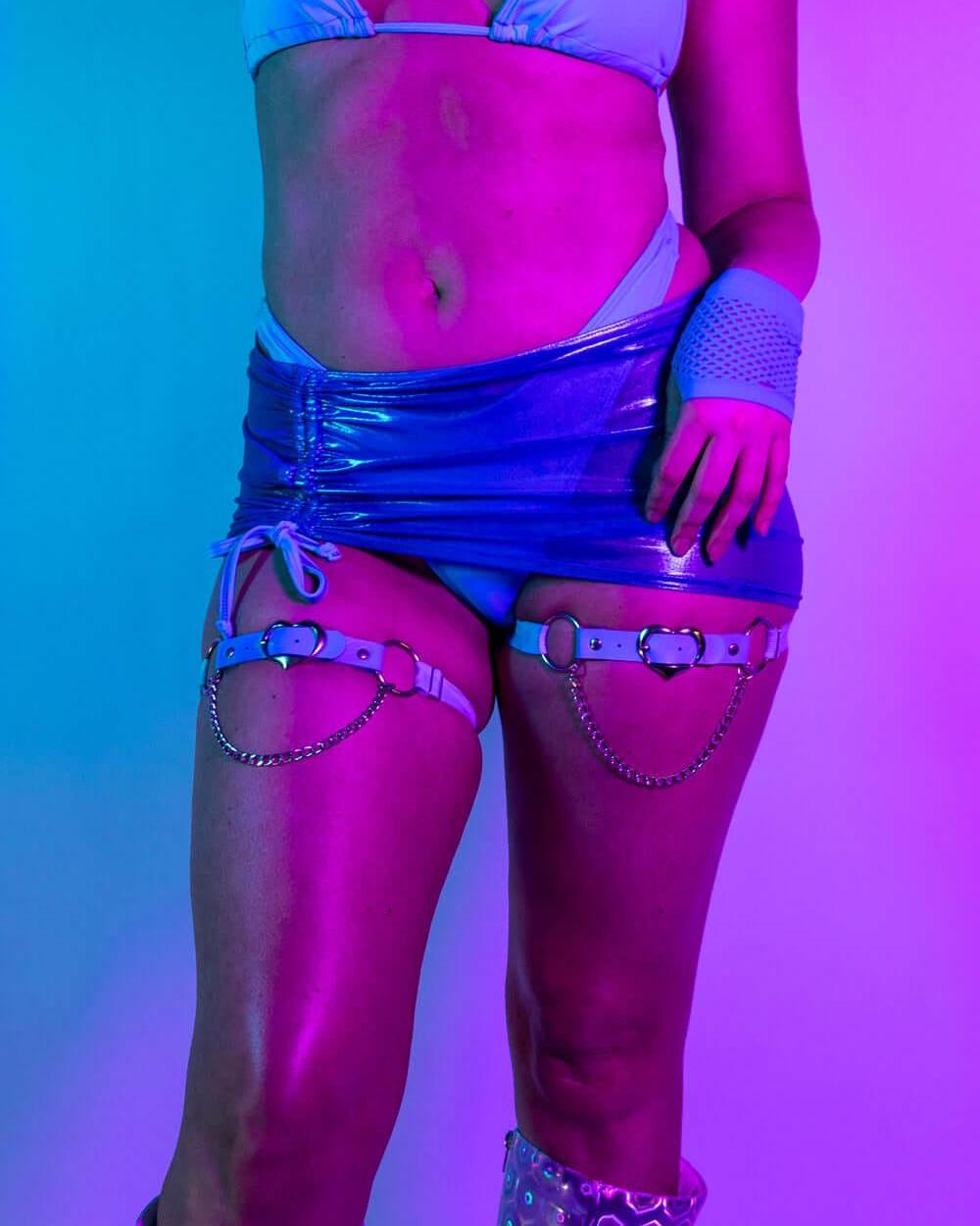Model wearing shimmering blue mesh cinch skirt and bikini, showcasing rave outfits under vibrant purple lighting.