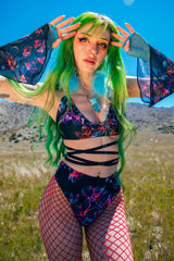 Vibrant portrait of a model with green and blue hair in a Freedom Rave Wear floral bodysuit and red fishnet tights, posing outdoors under clear skies.