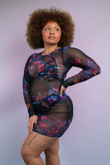 Model with curly hair in a Freedom Rave Wear floral mesh dress with sheer panels, looking up thoughtfully, against a soft purple backdrop.