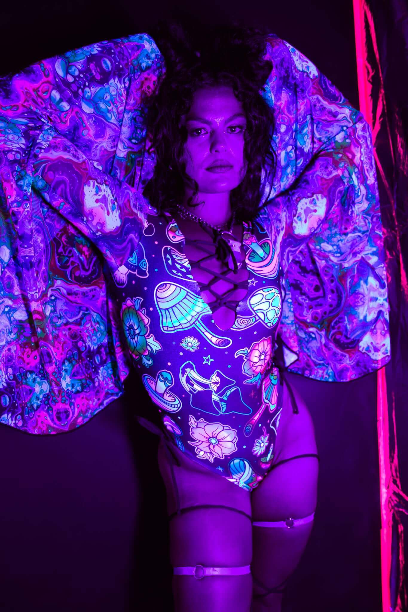 Model showcases Freedom Rave Wear's UV-reactive bodysuit featuring glowing mushroom and floral patterns under blacklight, paired with a matching shawl for a bold rave look.