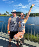 Festival couple in matching black and white optical illusion outfits by Freedom Rave Wear, standing by the water, embracing the sunny vibes."