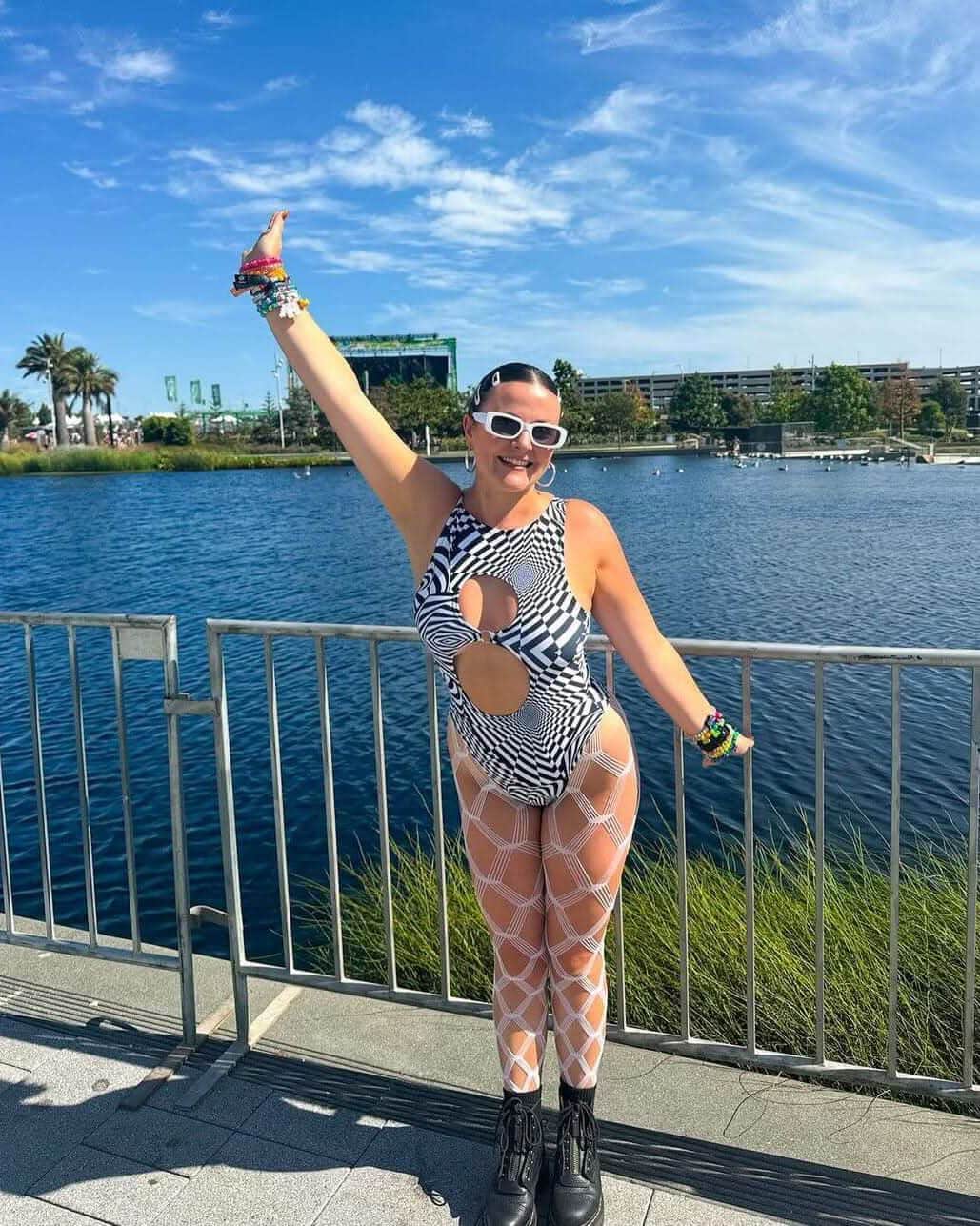 Woman in a Distortion Keyhole Bodysuit posing by water, showcasing rave outfit with cutouts and stylish accessories.