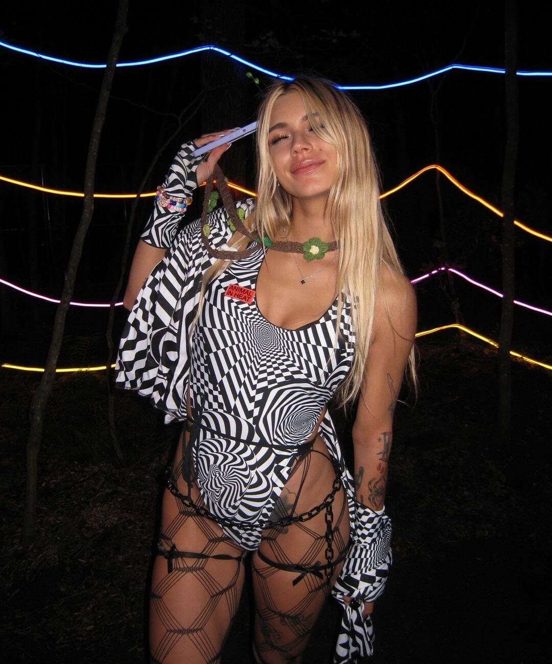 Model in a black and white optical illusion print rave bodysuit by Freedom Rave Wear, paired with chain accessories and mesh tights, under neon lights.