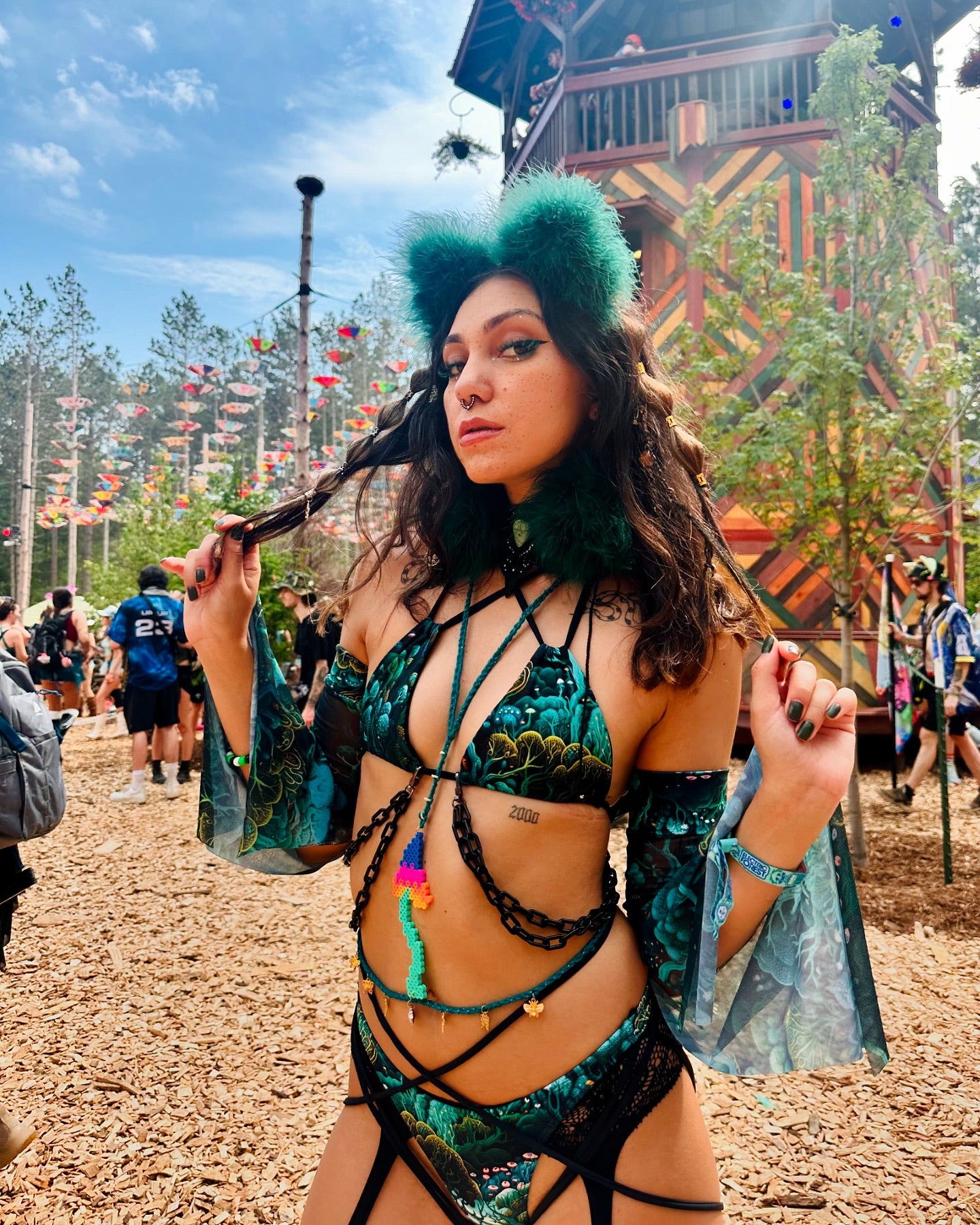 Festival-goer in a green, forest-themed rave outfit with flared sleeves and cat ears poses confidently at a vibrant, outdoor festival