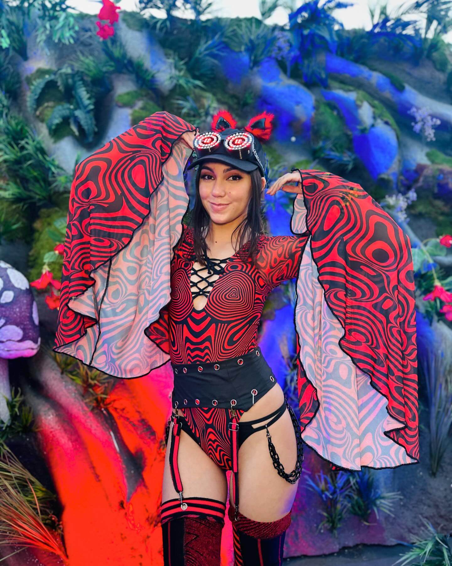 Rave fashionista in red and black patterned attire with LED goggles and kitty ears, posing in a whimsical festival environment