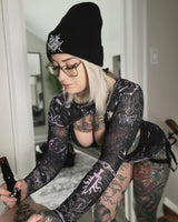 girl in a purple and black mesh rave outfit with tattoos style designs sits in her home drawing tattoo designs