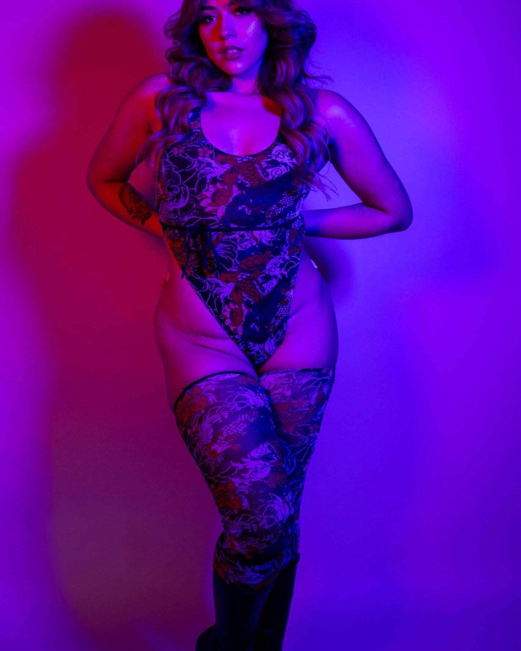 Neon-lit shot of the Good Fortune Mesh Temptation One Piece, highlighting its sheer design and bold tattoo-inspired artwork.