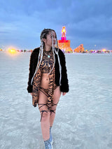 Adventurous raver in tribal bodysuit and faux fur coat stands on the Playa at dusk with the Burning Man effigy aglow in the background