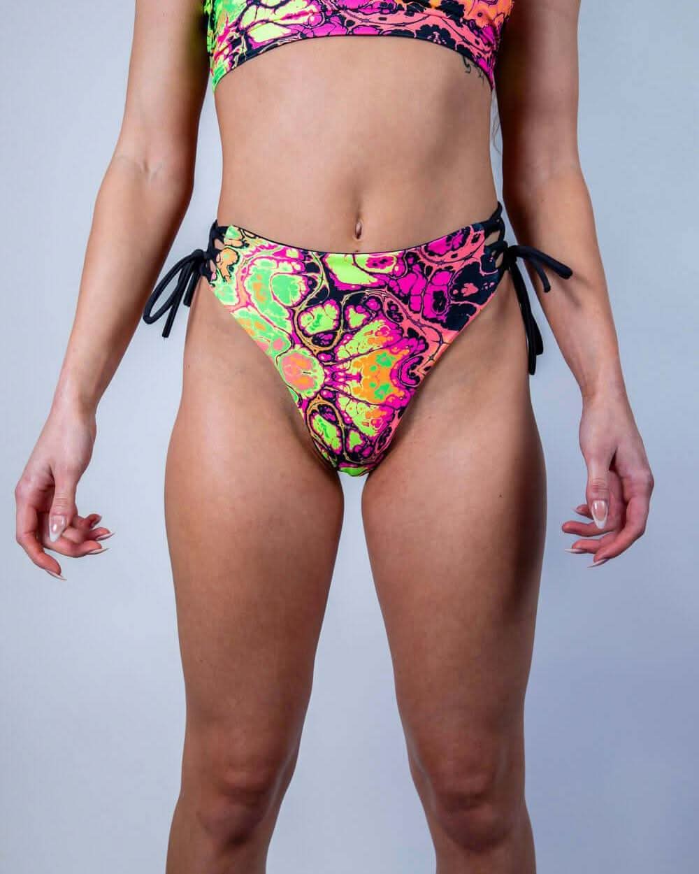 Model wearing Intensity Lace Up Brazilian Bottoms in vibrant patterns, ideal for rave outfits and stylish summer swimwear.